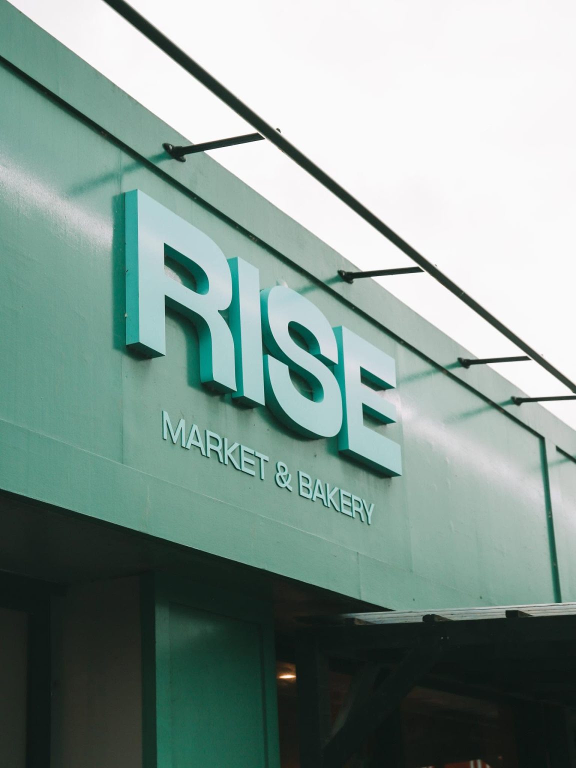 Exterior of Rise Market & Bakery, a teal-coloured food market and bakery with large signage reading “RISE”.