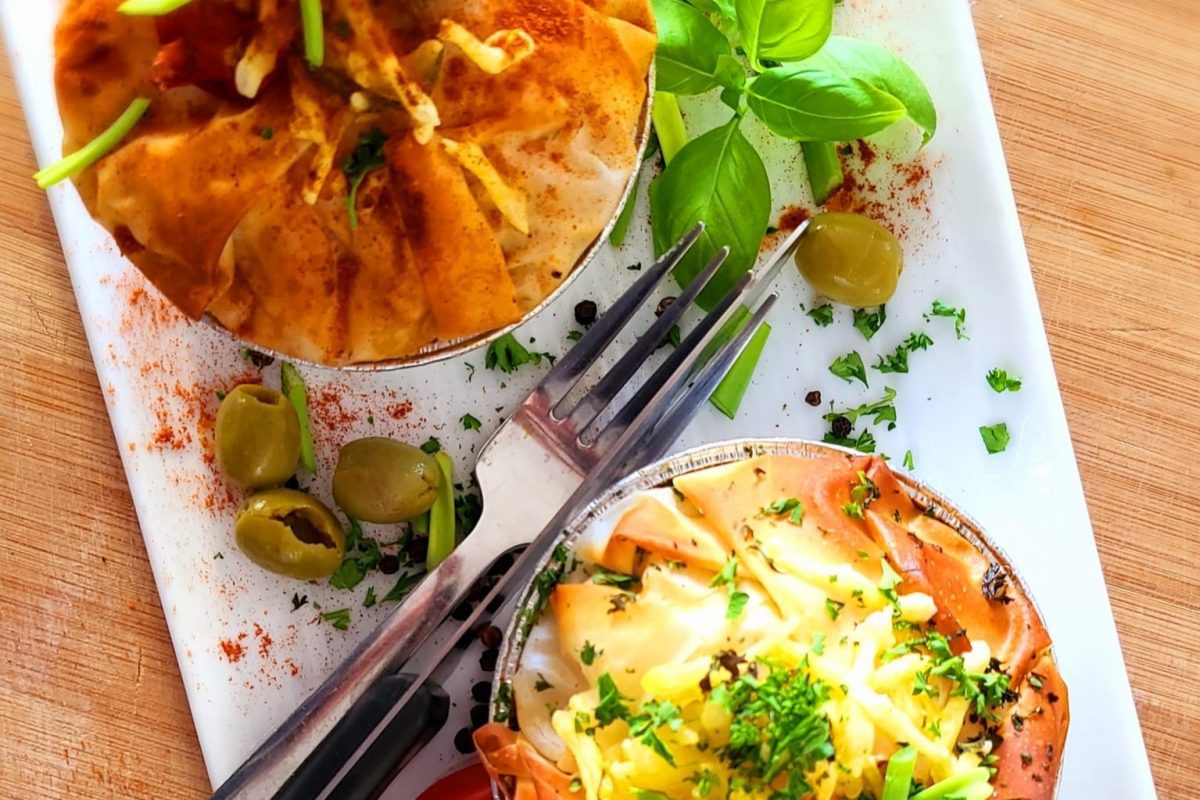 Two individual filo pastry pies on a white plate, garnished with chopped herbs, olives, and fresh basil leaves, with a fork placed between them.