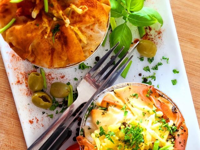 Two individual filo pastry pies on a white plate, garnished with chopped herbs, olives, and fresh basil leaves, with a fork placed between them.