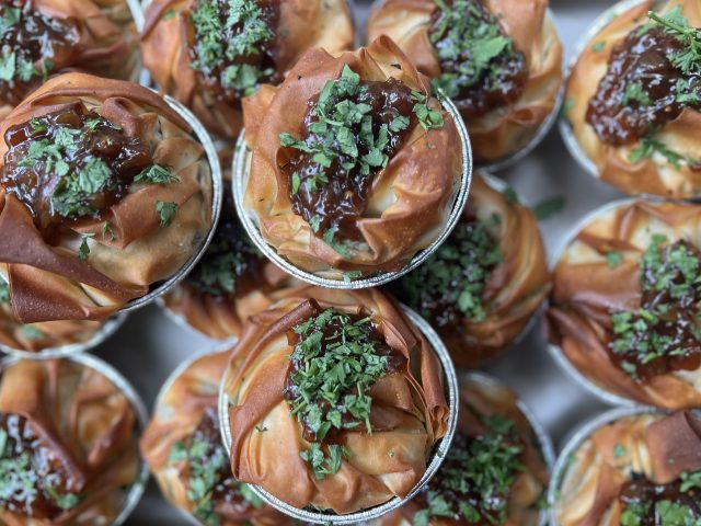 A close-up view of multiple filo pastry pies with golden, crispy layers, topped with a dark glaze and sprinkled with chopped fresh herbs.