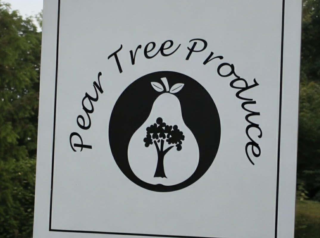 White hanging sign reading ‘Pear Tree Produce’ with a black pear-shaped logo containing a tree, suspended from a wooden beam outdoors with greenery behind.