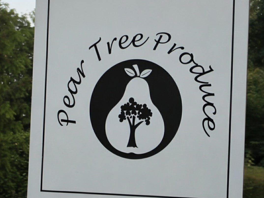 White hanging sign reading ‘Pear Tree Produce’ with a black pear-shaped logo containing a tree, suspended from a wooden beam outdoors with greenery behind.