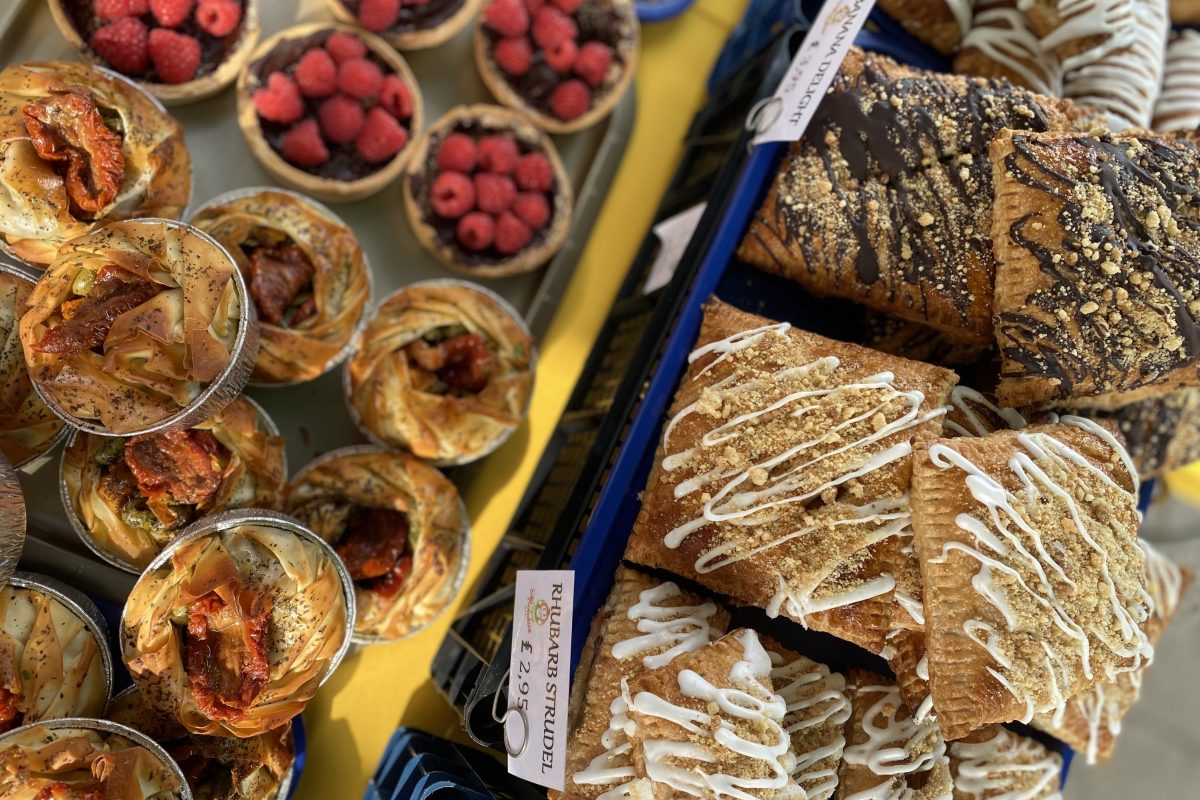 A market stall display featuring a variety of pastries, including raspberry tarts, filo pies with roasted tomatoes, and rectangular pastries drizzled with icing.