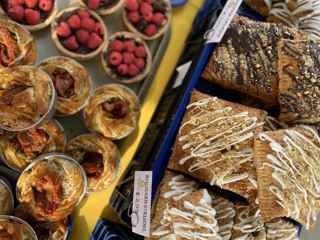 A market stall display featuring a variety of pastries, including raspberry tarts, filo pies with roasted tomatoes, and rectangular pastries drizzled with icing.