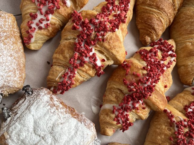An assortment of pastries including croissants topped with icing and dried raspberry pieces, alongside pastries dusted with powdered sugar.