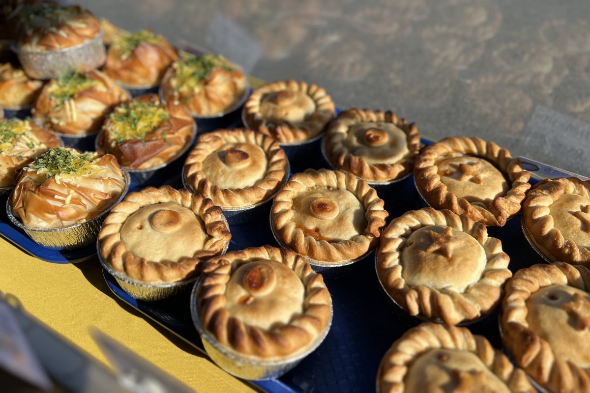 A market stall display of traditional round pies with crimped edges and some filo pastry pies in the background, all arranged on blue trays in natural sunlight.