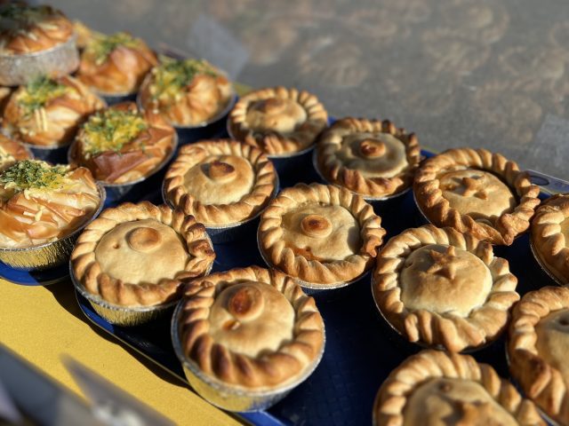 A market stall display of traditional round pies with crimped edges and some filo pastry pies in the background, all arranged on blue trays in natural sunlight.