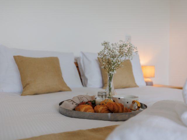 Light and airy bedroom with a neatly made double bed, soft neutral cushions, bedside lamps and a breakfast tray with croissants and coffee.