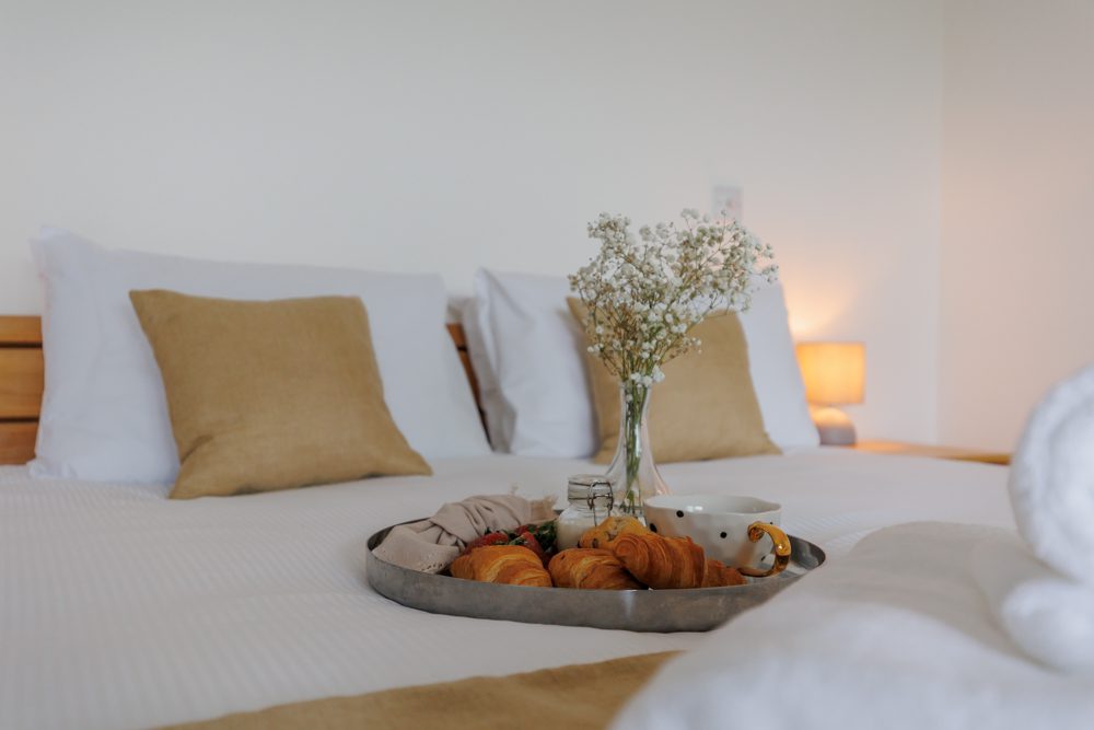 Light and airy bedroom with a neatly made double bed, soft neutral cushions, bedside lamps and a breakfast tray with croissants and coffee.