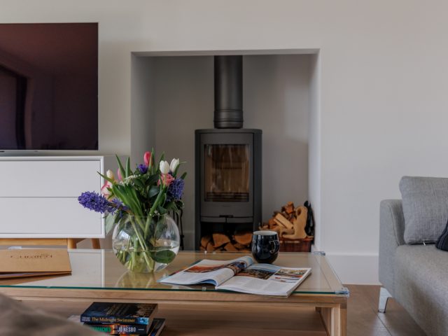 Stylish living room featuring a log burner set into a wall alcove, a glass coffee table with flowers and magazines, and modern seating.