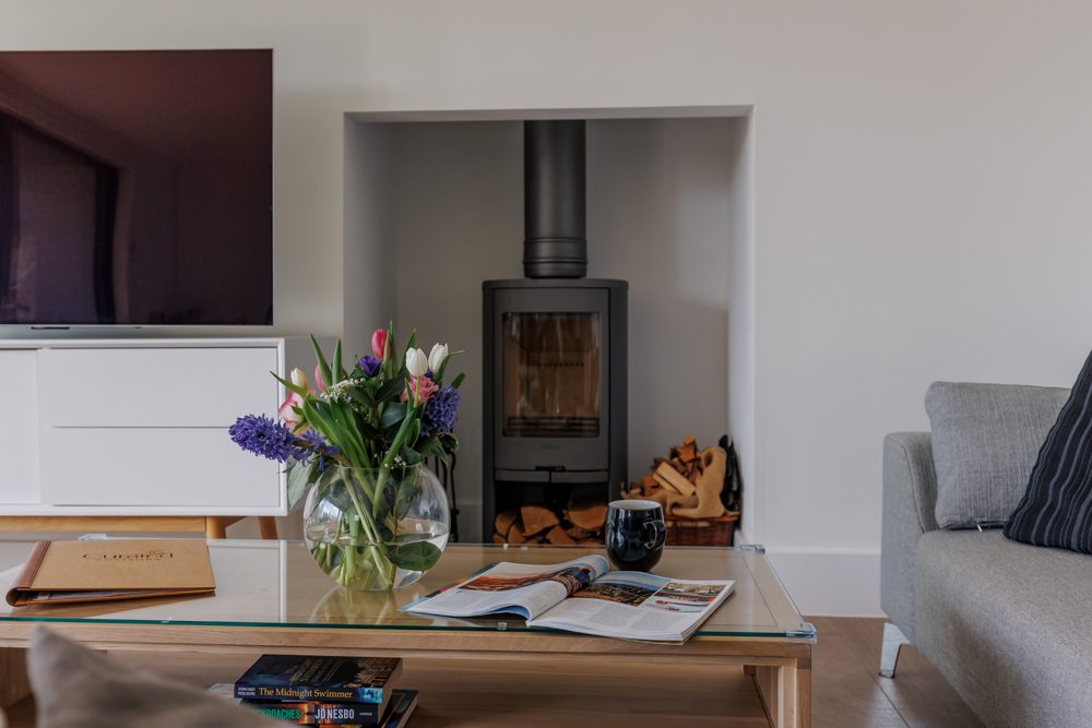 Stylish living room featuring a log burner set into a wall alcove, a glass coffee table with flowers and magazines, and modern seating.
