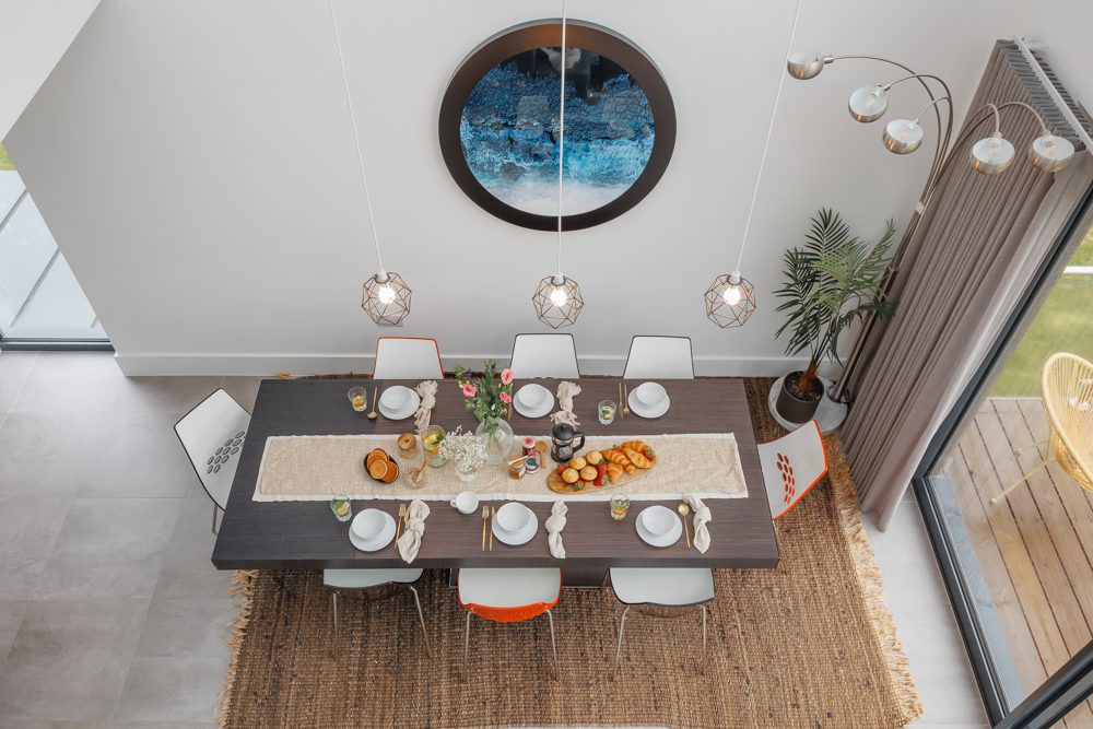 Overhead view of a modern dining table set for breakfast, with plates, pastries, flowers and pendant lights in a bright, contemporary holiday cottage dining space.