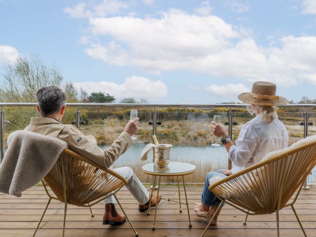 Two guests seated on a balcony overlooking calm water and countryside, raising glasses on a small table with outdoor chairs and open views.