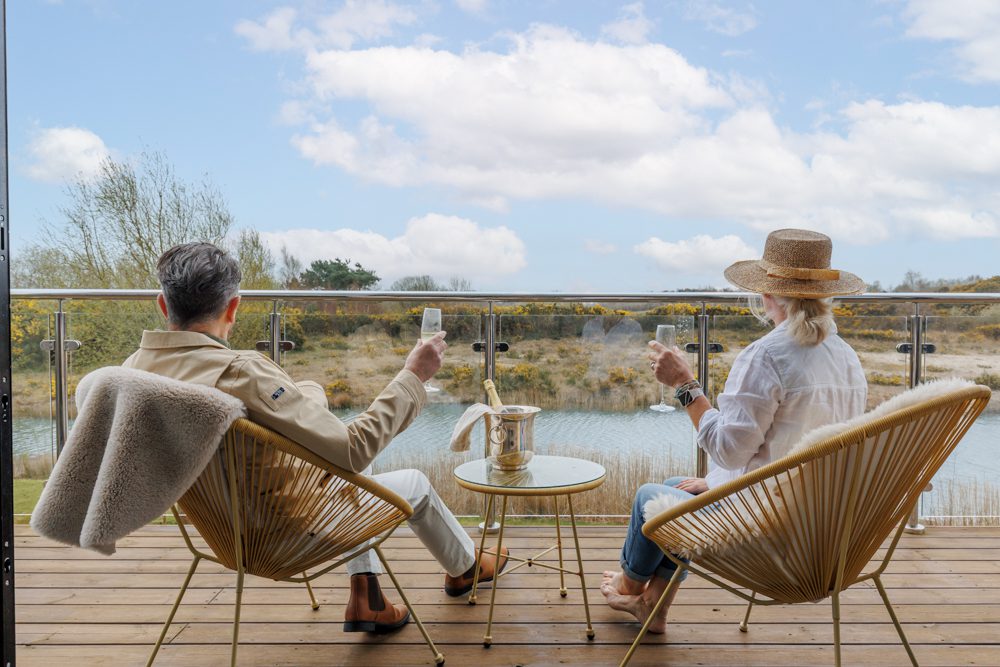 Two guests seated on a balcony overlooking calm water and countryside, raising glasses on a small table with outdoor chairs and open views.