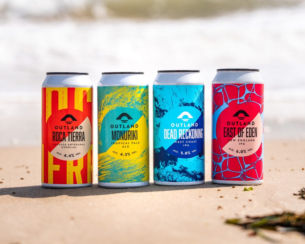 Four colourful Outland craft beer cans standing upright on a sandy beach, each with bold, bright label designs labelled “Roca Tierra,” “Monuriki,” “Dead Reckoning,” and “East of Eden,” with a blurred shoreline in the background.