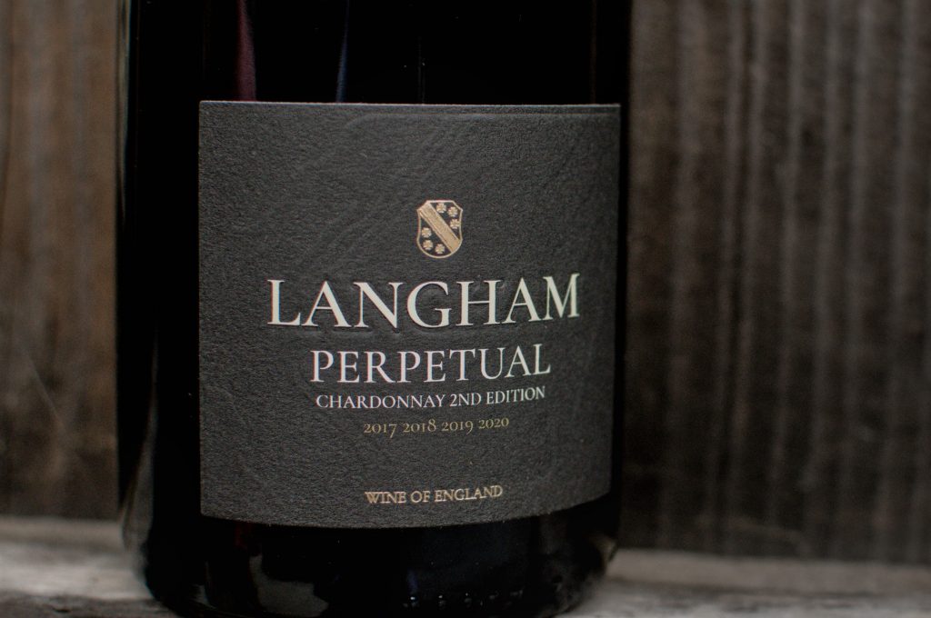 Close-up of a Langham Wine Estate “Perpetual” Chardonnay Second Edition sparkling wine bottle, showing a dark label with white lettering and vintage years 2017–2020, set against a rustic wooden background.