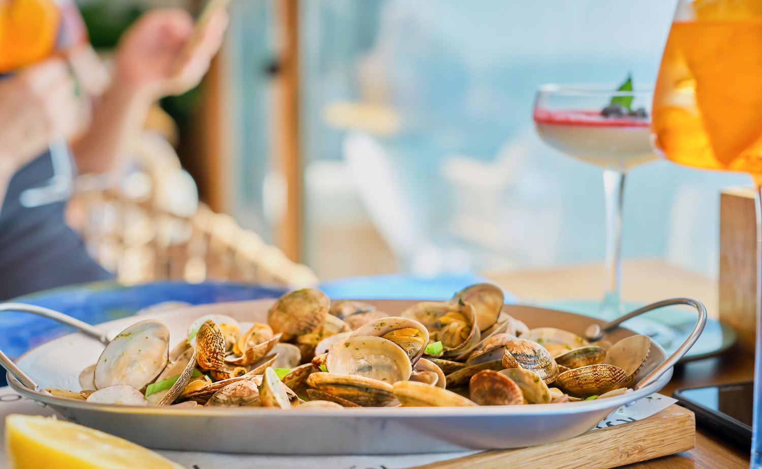 Pan of cooked clams with herbs and lemon on a table, with drinks and a blurred outdoor dining scene in the background.