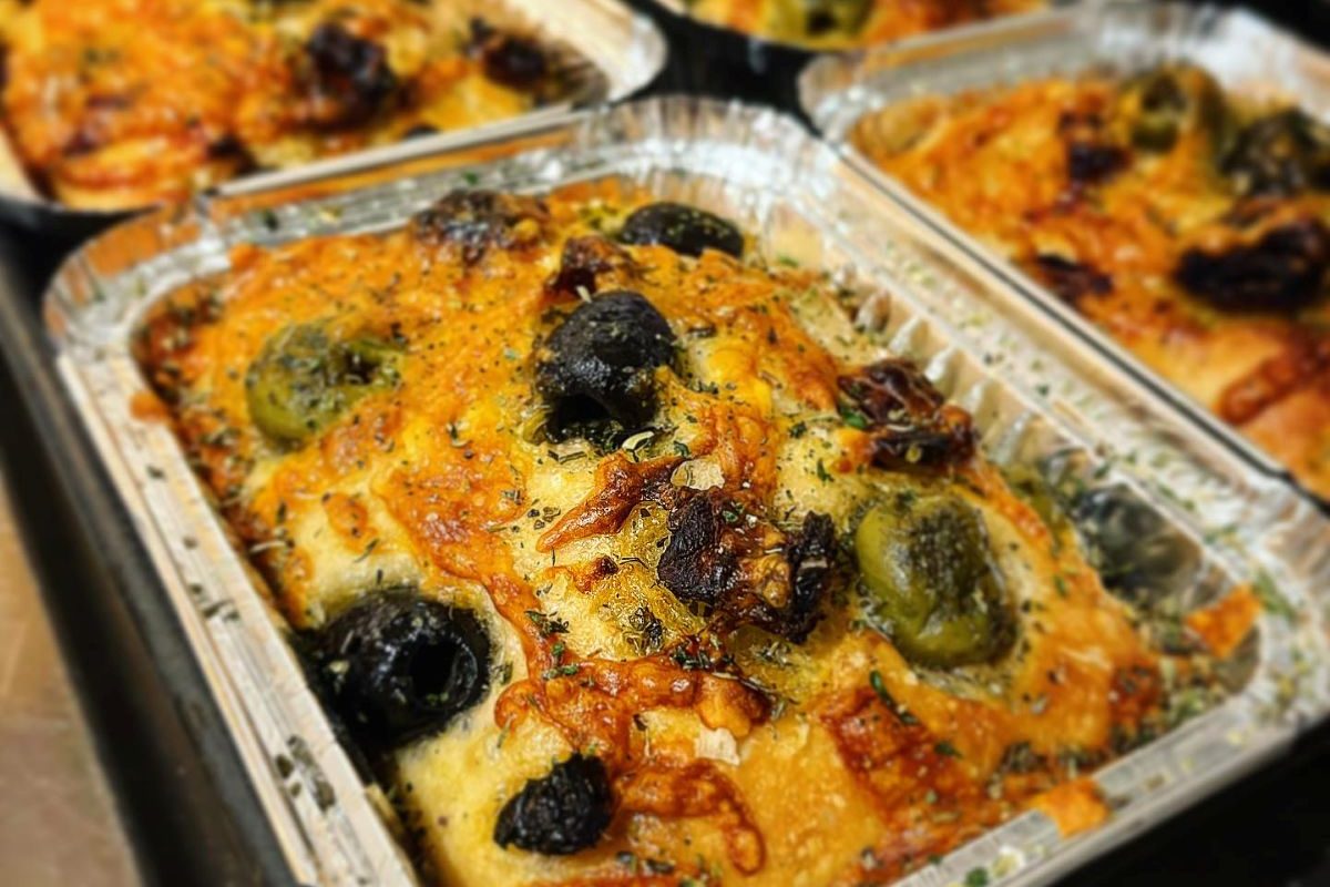 Freshly baked focaccia topped with green and black olives, melted cheese and herbs, displayed in bakery trays ready for sale.