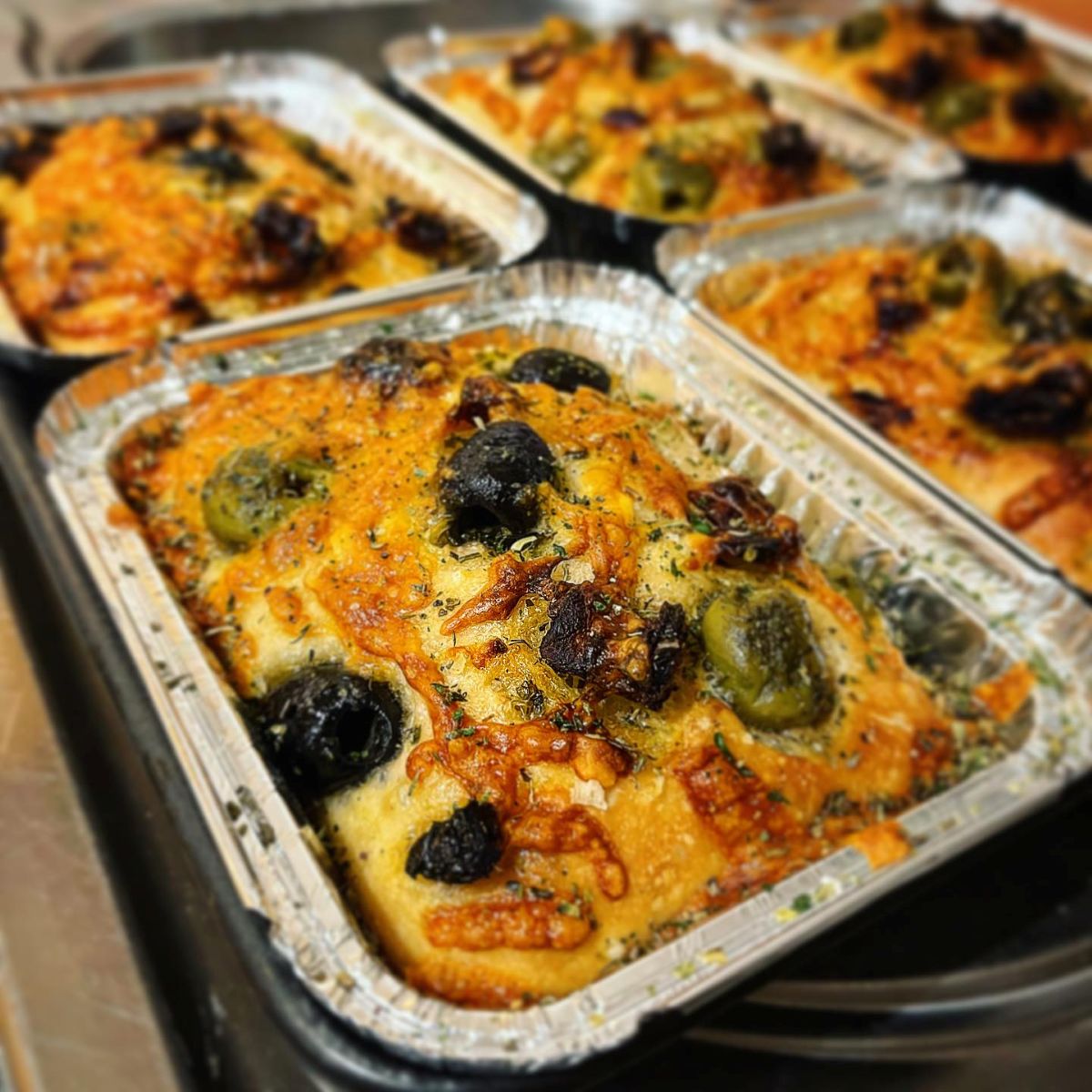 Freshly baked focaccia topped with green and black olives, melted cheese and herbs, displayed in bakery trays ready for sale.