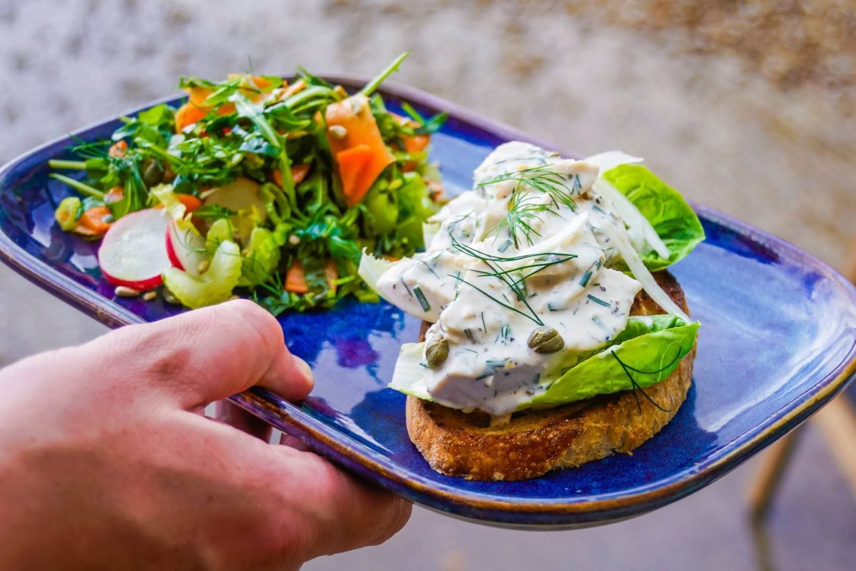 A colourful brunch dish served on a deep blue plate, featuring creamy herb-coated chicken on toasted artisan bread with crisp lettuce leaves, capers and fresh dill. Served alongside a vibrant mixed salad of leafy greens, sliced radish, carrot ribbons and seeds. The plate is being held outdoors, highlighting the fresh, homemade style of food served at Cherries at Abbey Farm.