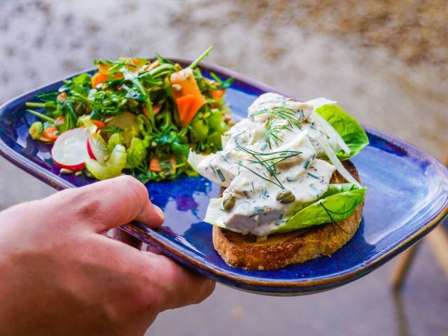 A colourful brunch dish served on a deep blue plate, featuring creamy herb-coated chicken on toasted artisan bread with crisp lettuce leaves, capers and fresh dill. Served alongside a vibrant mixed salad of leafy greens, sliced radish, carrot ribbons and seeds. The plate is being held outdoors, highlighting the fresh, homemade style of food served at Cherries at Abbey Farm.