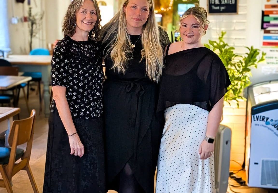Three members of the Cherries at Abbey Farm team standing together inside the café. The bright and welcoming interior features wooden furniture, decorative lighting and a relaxed dining space that reflects the venue's friendly, community-focused atmosphere.