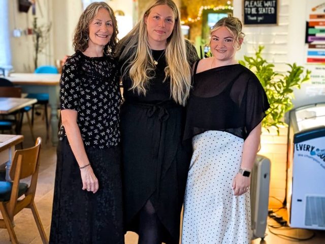 Three members of the Cherries at Abbey Farm team standing together inside the café. The bright and welcoming interior features wooden furniture, decorative lighting and a relaxed dining space that reflects the venue's friendly, community-focused atmosphere.