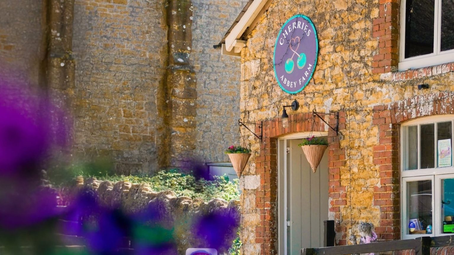 Visitors gather outside a historic stone barn at Abbey Farm in Abbotsbury during a summer community event. Colourful bunting is strung across the foreground, while market-goers and families explore the site and enjoy activities near the barn entrance. Wildflowers and greenery frame the scene, showcasing the relaxed countryside atmosphere and community spirit of a rural Dorset farm event.