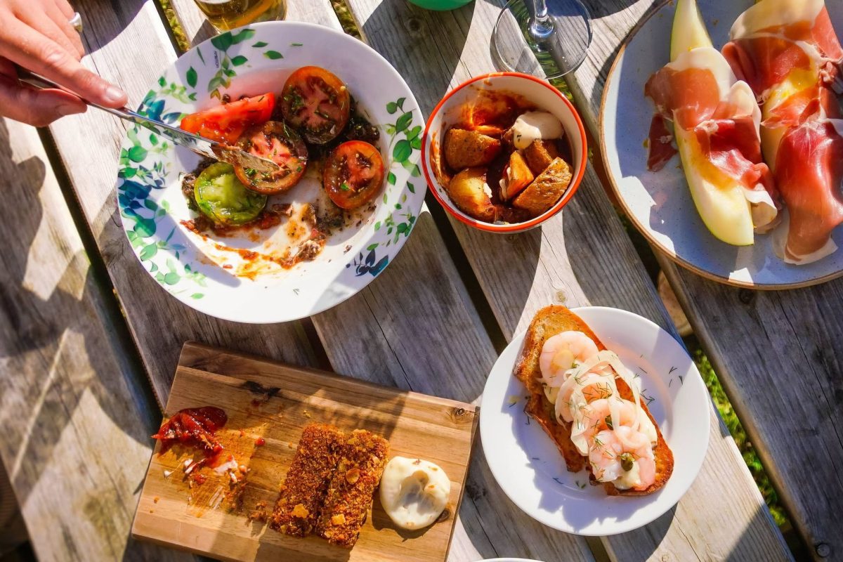 An overhead view of a table filled with Mediterranean-inspired sharing dishes enjoyed outdoors in the sunshine. Plates include grilled tomatoes, toasted bread topped with prawns, cured ham served with melon, crispy bites with dipping sauce, olives and roasted potatoes, accompanied by drinks and glasses on a rustic wooden table.