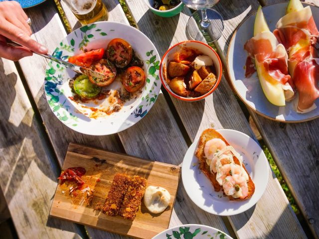 An overhead view of a table filled with Mediterranean-inspired sharing dishes enjoyed outdoors in the sunshine. Plates include grilled tomatoes, toasted bread topped with prawns, cured ham served with melon, crispy bites with dipping sauce, olives and roasted potatoes, accompanied by drinks and glasses on a rustic wooden table.
