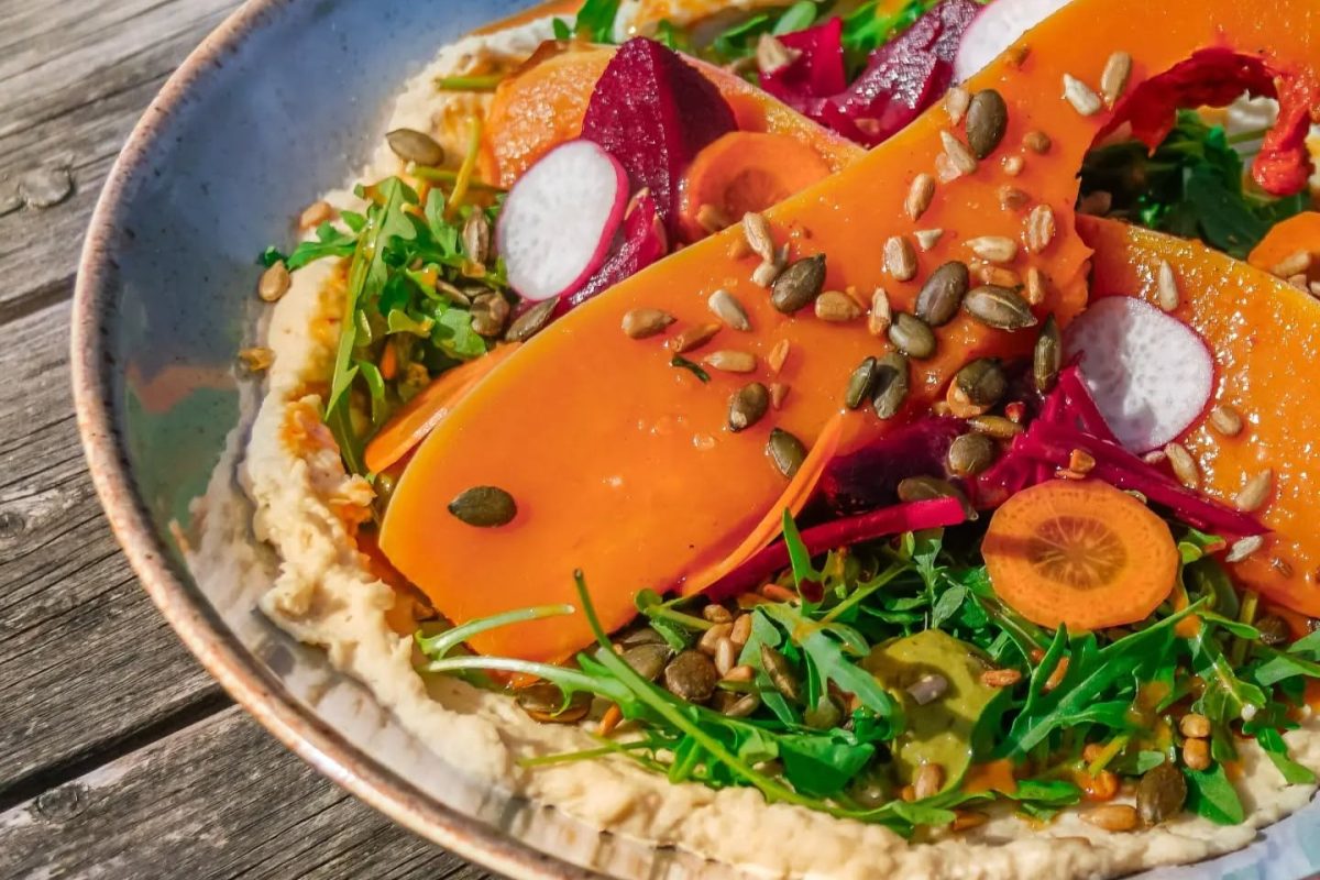 A colourful roasted squash salad served in a shallow bowl, featuring ribbons of harissa honey-roasted squash layered over creamy hummus. The dish is topped with rocket leaves, sliced radishes, pickled vegetables, carrot slices and a scattering of pumpkin and sunflower seeds, creating a vibrant and seasonal lunch dish.