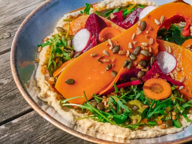 A colourful roasted squash salad served in a shallow bowl, featuring ribbons of harissa honey-roasted squash layered over creamy hummus. The dish is topped with rocket leaves, sliced radishes, pickled vegetables, carrot slices and a scattering of pumpkin and sunflower seeds, creating a vibrant and seasonal lunch dish.